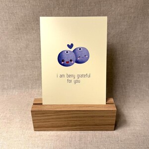 May include: A greeting card with a light yellow background, featuring two cartoon blueberries with smiling faces and a small blue heart. The text "i am berry grateful for you" is below the blueberries. The card is displayed in a wooden block.
