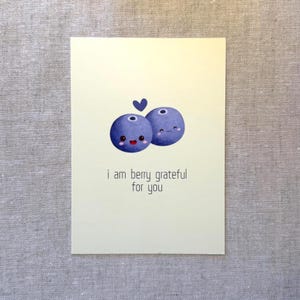 May include: A greeting card with a light yellow background features two cartoon blueberries with smiling faces and a small blue heart above them. The text "i am berry grateful for you" is printed below the blueberries.