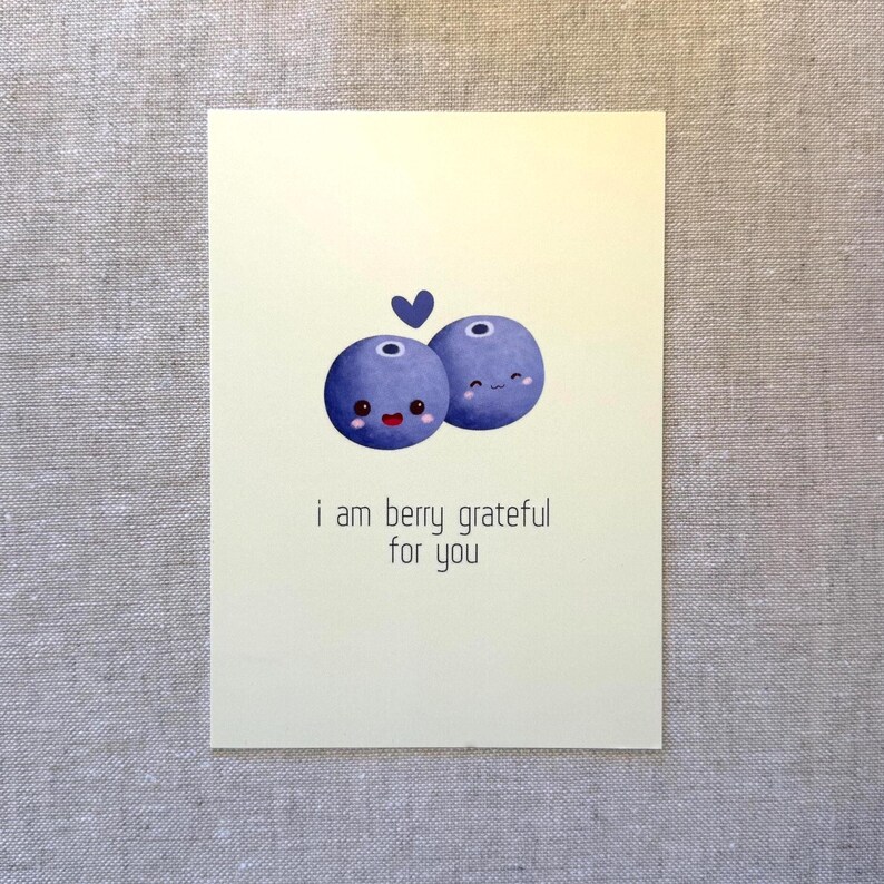 May include: A greeting card with a light yellow background features two cartoon blueberries with smiling faces and a small blue heart above them. The text "i am berry grateful for you" is printed below the blueberries.