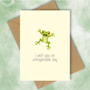 Könnte beinhalten: Grußkarte mit hellgelbem Hintergrund, mit einer grünen Cartoon-Frosch mit gelben Augen und einem lächelnden Mund. Der Text "i wish you an unforgettable day" steht darunter. Die Karte liegt auf einem braunen Umschlag.