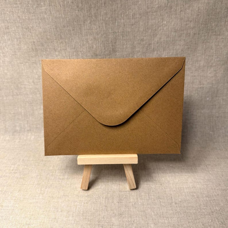 May include: A brown paper envelope, displayed on a small wooden easel. The envelope is rectangular with a triangular flap. The background is a neutral, textured fabric. The envelope is a warm, earthy tone.