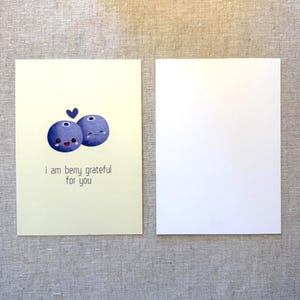 May include: Two greeting cards. One card has a light yellow background with a cartoon blueberry design and the text "i am berry grateful for you". The other card is blank. The blueberries are blue with smiling faces.