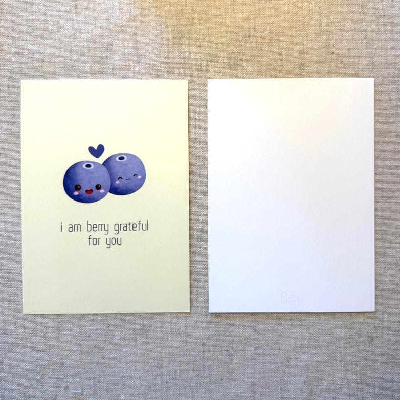 May include: Two greeting cards. One card has a light yellow background with a cartoon blueberry design and the text "i am berry grateful for you". The other card is blank. The blueberries are blue with smiling faces.