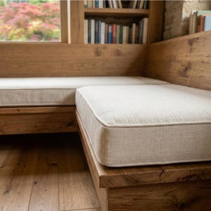 May include: A custom L-shaped wooden bench featuring a natural oak finish and thick, light beige fabric cushions. The bench is built into a corner with integrated shelving filled with books, positioned next to a window with a view of trees.