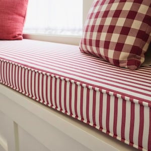 May include: A window seat cushion and pillows with a red and white striped pattern. The pillows have a red and white checkered pattern. The seat is made of a soft, comfortable material.
