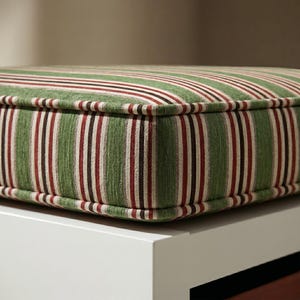 Peut inclure: Gros plan sur un coussin rectangulaire à rayures vertes et blanches, avec des rayures bordeaux et noires. Le coussin a une bordure épaisse et rembourrée et est posé sur une surface blanche.