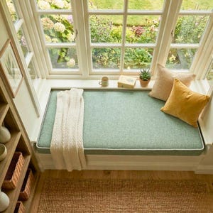 May include: A window seat with a light green cushion and white trim. A cream-coloured throw blanket is draped over the seat. Two pillows, one tan and one yellow, rest on the cushion. A small potted plant, a mug, and a book sit on the windowsill.