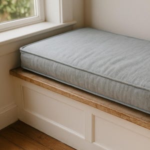 May include: A light gray rectangular cushion with a contrasting border sits atop a wooden bench. The bench is built into a white paneled structure, near a window. The cushion appears to be made of a textured fabric.
