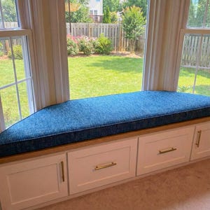 May include: A custom-built window seat with a blue cushion and white cabinets. The seat features a wooden base and gold-colored hardware. The window overlooks a backyard with a green lawn and a wooden fence.