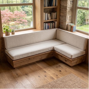 May include: An L-shaped corner bench featuring a natural wood frame and thick, off-white upholstered cushions. The seating unit is positioned against a wooden bookshelf and large windows, providing a structured, neutral-toned furniture piece.