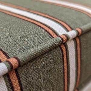 May include: A textured fabric cushion featuring a striped pattern in shades of olive green, white, and burnt orange. The cushion has a prominent piped edge detail that runs along the seam, highlighting the woven construction of the textile.