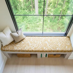 May include: A window seat with a cream and gold floral patterned cushion. Two white pillows with pom-pom trim and a tan pillow rest on the seat. Two woven baskets and a stack of books are stored below the seat.