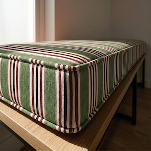 Peut inclure: Banc avec coussin à rayures vertes, rouges, blanches et noires. Le coussin est rectangulaire et possède une bordure épaisse et rembourrée. Le banc a un cadre en bois clair et des pieds en métal noir. Les rayures du coussin sont verticales.