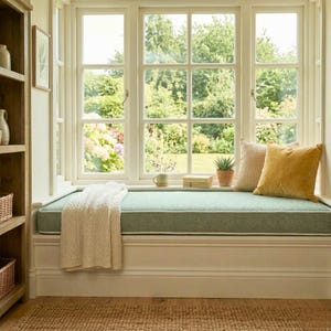 May include: A window seat with a light green cushion and white trim. A cream-coloured throw blanket is draped over the seat. Two pillows, one striped and one mustard yellow, rest against the window. A small potted plant and a mug are on the seat.