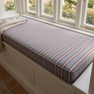 May include: A rectangular window seat cushion featuring a blue, cream, and burgundy striped pattern. The cushion is tailored with piped edges and rests on a white wooden base. A folded, textured cream-colored throw blanket lies on the left side.
