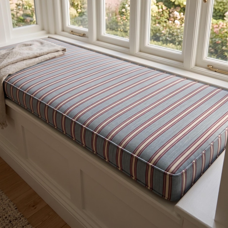 May include: A rectangular window seat cushion featuring a blue, cream, and burgundy striped pattern. The cushion is tailored with piped edges and rests on a white wooden base. A folded, textured cream-colored throw blanket lies on the left side.