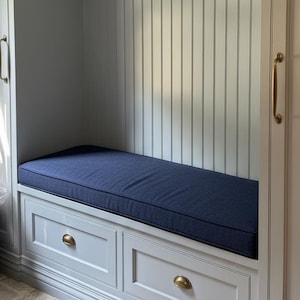 May include: A built-in bench with a navy blue cushion. The bench has two drawers with brass-colored hardware. The surrounding walls are painted light gray with vertical paneling. The overall design is clean and modern.