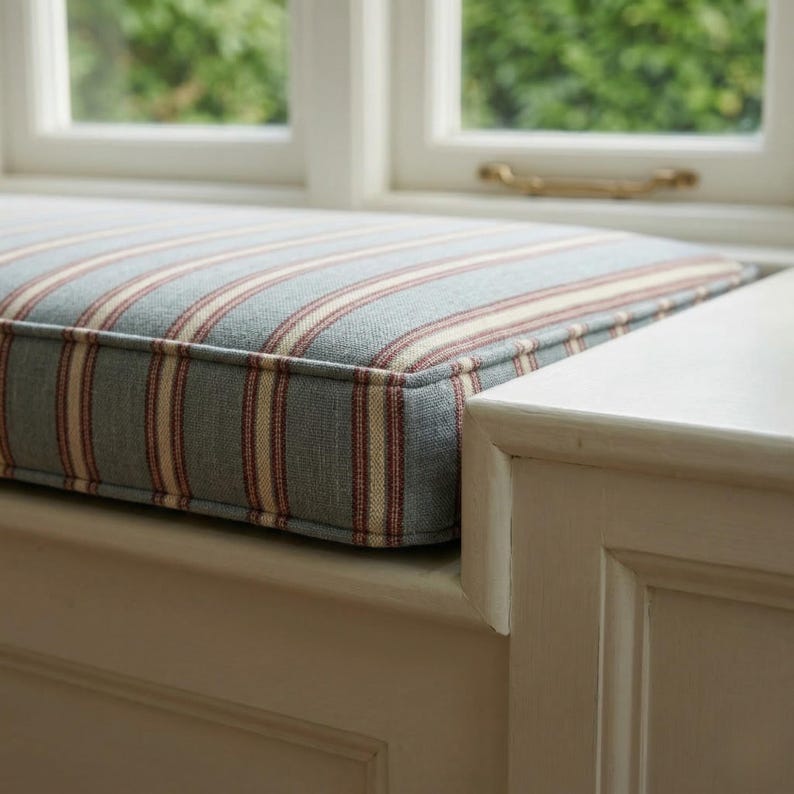 May include: A rectangular window seat cushion featuring a woven fabric with a pattern of vertical stripes in muted blue, cream, and thin dark red lines. The cushion has piped edges and is positioned on a white wooden base near a window frame.