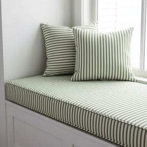 Peut inclure: Un coussin de banquette et deux coussins assortis. Les coussins présentent un motif à rayures vertes et blanches. La banquette est encastrée dans un cadre blanc, avec une fenêtre en arrière-plan.
