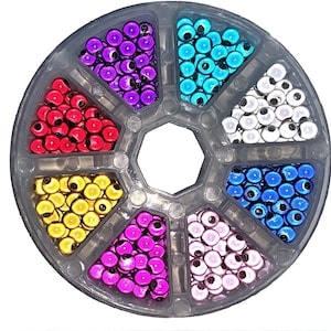 Puede incluir: Un organizador de plástico transparente lleno de cuentas de manualidades de colores. Las cuentas están dispuestas en siete secciones, cada una con un color diferente: rojo, morado, turquesa, blanco, azul, rosa y amarillo. Cada cuenta tiene un centro negro.