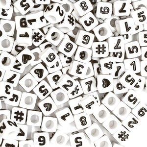 May include: A collection of white plastic cube beads with black numbers and symbols, including numbers 1 through 9, the pound sign, and a heart symbol.
