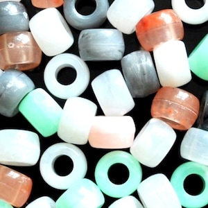 May include: A selection of beads in a variety of colours and forms. The beads include translucent orange, white, grey, and green barrel-shaped beads, alongside white and green donut-shaped beads. The beads are scattered on a black surface.