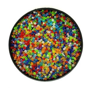 Op de afbeelding: Een zwarte ronde container gevuld met een levendig assortiment kleine, kleurrijke kralen. De kralen zijn een mix van rood, oranje, geel, groen, blauw en paars, waardoor een regenboogeffect ontstaat. De kralen zijn klein en lijken van glas of plastic te zijn.