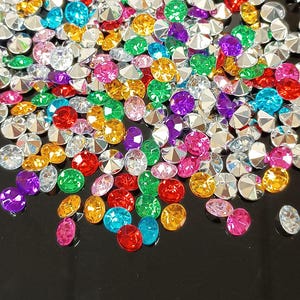 Könnte beinhalten: Eine Ansammlung bunter Strasssteine in verschiedenen Formen und Größen. Die Edelsteine sind in den Farbtönen Rot, Grün, Gelb, Lila, Rosa, Blau und Klar auf einer dunklen Oberfläche verstreut. Diese funkelnden Verzierungen sind ideal für Bastelprojekte.