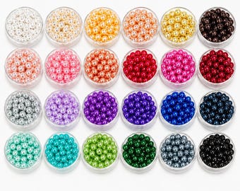 May include: A collection of small, round beads in various colors, including white, gold, orange, pink, red, purple, blue, green, and black. Each color is contained in a clear, circular container, creating a visually appealing display of craft supplies.