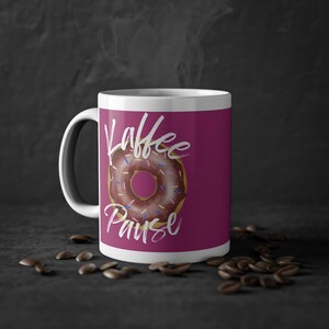 Könnte beinhalten: Weiße Keramiktasse mit rosa Hintergrund und einem Donut mit Streuseln. Der Text "Kaffee Pause" ist in Weiß auf die Tasse geschrieben.