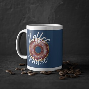 Könnte beinhalten: Weiße Keramiktasse mit blauem Hintergrund und einem Donut mit rosa Zuckerguss und bunten Streuseln. Der Text "Kaffee Pause" ist in weißer Kursivschrift auf der Tasse geschrieben.