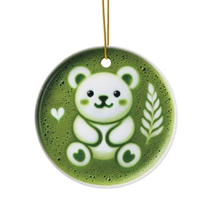 以下が含まれることがあります： 緑色の背景に白いクマのデザインが施された丸いセラミックオーナメント。クマは可愛らしい顔をしており、ハートと葉のデザインに囲まれています。吊り下げ用の金色の紐が付いています。