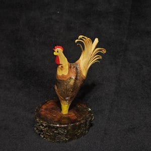 May include: A handcrafted wooden rooster sculpture. The rooster features a red comb and wattle, yellow beak, and a detailed, fanned tail. It is mounted on a small, round, wooden base, showcasing the natural wood grain and textures.
