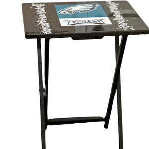Peut inclure: Table pliante noire avec un logo Eagles noir et blanc sur le dessus. La table a un cadre en métal noir.