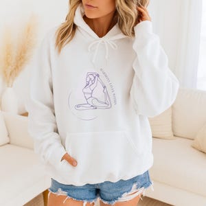 Könnte beinhalten: Weißer Kapuzenpullover mit Fronttasche und Kordelzug. Er zeigt eine lila Strichzeichnung einer Person in einer Yoga-Pose mit dem Text "HARMONY LIVES WITHIN". Das Design ist mittig auf der Vorderseite platziert.
