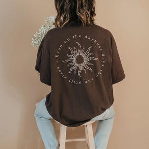 Könnte beinhalten: Ein braunes T-Shirt mit einem kreisförmigen Sonnengrafik und dem Text "Even on the darkest days the sun still rises". Die Person trägt eine hellblaue Jeans und sitzt auf einem Holzschemel.