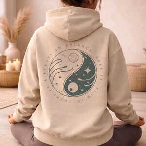 Könnte beinhalten: Beige Kapuzenpullover mit Kapuze und einem kreisförmigen Aufdruck auf dem Rücken. Das Design ist ein Yin-Yang-Design in Türkis und Weiß, mit den Worten "A BEAUTY IN BALANCE. SEEK IT. FIND IT. THERE IS" am Rand. Die Person trägt graue Leggings.