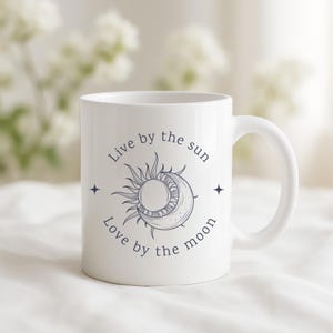 May include: White ceramic mug with a celestial design featuring a sun and moon illustration, accompanied by the text "Live by the sun, Love by the moon." The mug has a curved handle and is suitable for hot or cold beverages.