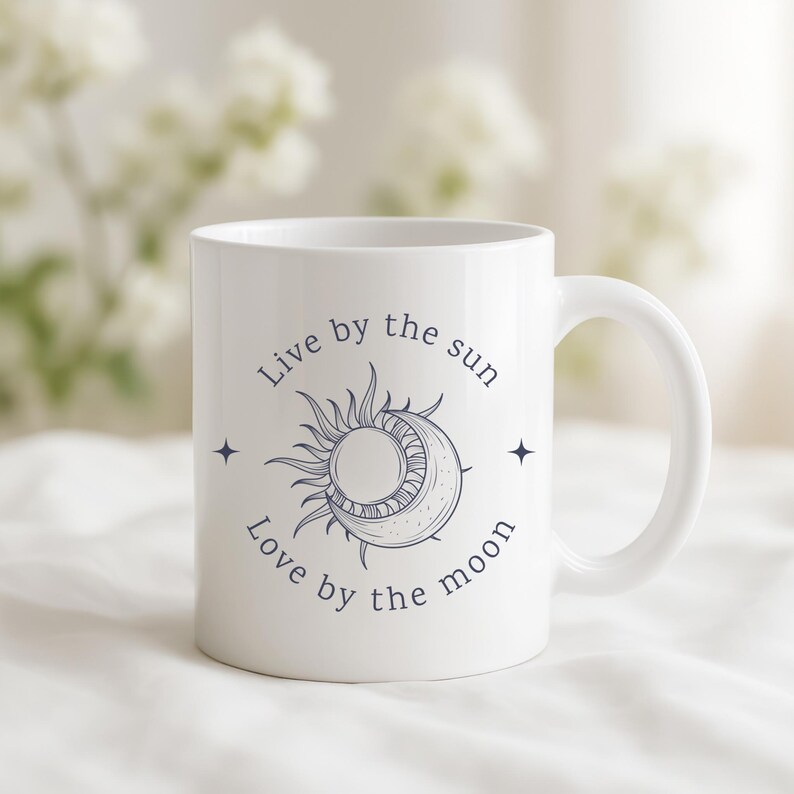 May include: White ceramic mug with a celestial design featuring a sun and moon illustration, accompanied by the text "Live by the sun, Love by the moon." The mug has a curved handle and is suitable for hot or cold beverages.