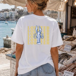 Könnte beinhalten: Ein weißes kurzärmeliges T-Shirt mit einer blauen Strichzeichnung eines Hummers auf der Rückseite. Das Wort LOBSTER ist in gelben Großbuchstaben wiederholt in einem vertikalen Säulenmuster hinter der Hummergrafik aufgedruckt.