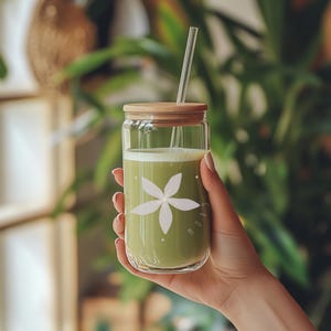 Könnte beinhalten: Ein klares Glas mit einem Holzdeckel und einem durchsichtigen Strohhalm, gefüllt mit einem grünen Getränk. Das Glas hat ein weißes Blumendesign und das Wort "GROW". Das Glas wird von einer Person gehalten.