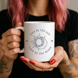 May include: White ceramic mug featuring a celestial design with a sun and moon illustration in navy blue. The mug has the text "Live by the sun, Love by the moon" in navy blue. The mug is held by a person.