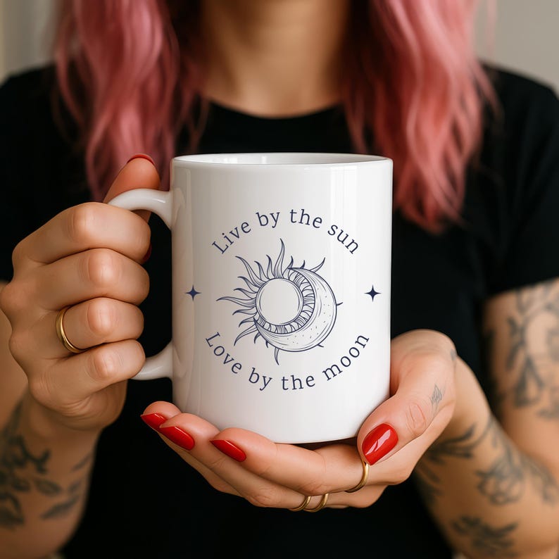 May include: White ceramic mug featuring a celestial design with a sun and moon illustration in navy blue. The mug has the text "Live by the sun, Love by the moon" in navy blue. The mug is held by a person.