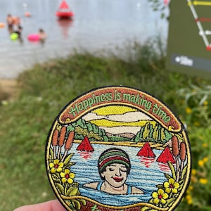 May include: A circular embroidered patch featuring a person swimming in a lake with cattails and yellow flowers on the sides. The text "Happiness is making time to swim" is stitched in green along the top and bottom borders of the design.