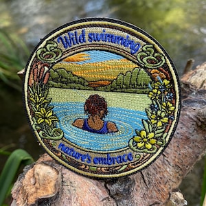 May include: A circular embroidered patch featuring a person swimming in a blue lake surrounded by green hills and yellow flowers. The text "Wild swimming" is at the top and "nature's embrace" is at the bottom in blue thread on a black border.
