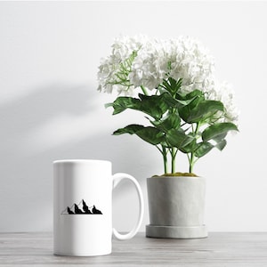 May include: White ceramic mug with a black mountain range design. The mug is sitting on a wooden table next to a potted plant with white flowers.