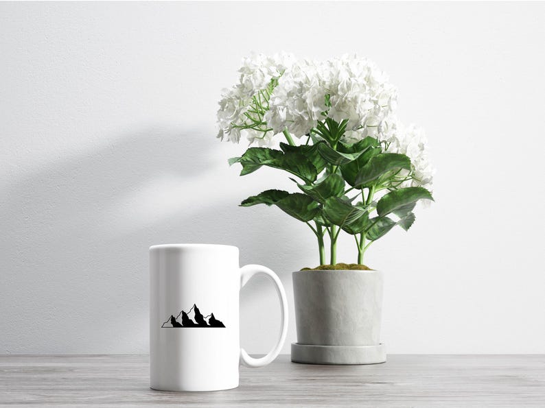 May include: White ceramic mug with a black mountain range design. The mug is sitting on a wooden table next to a potted plant with white flowers.