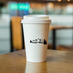 May include: White to-go coffee cup with a black mountain range and trees design. The cup is sitting on a wooden table.