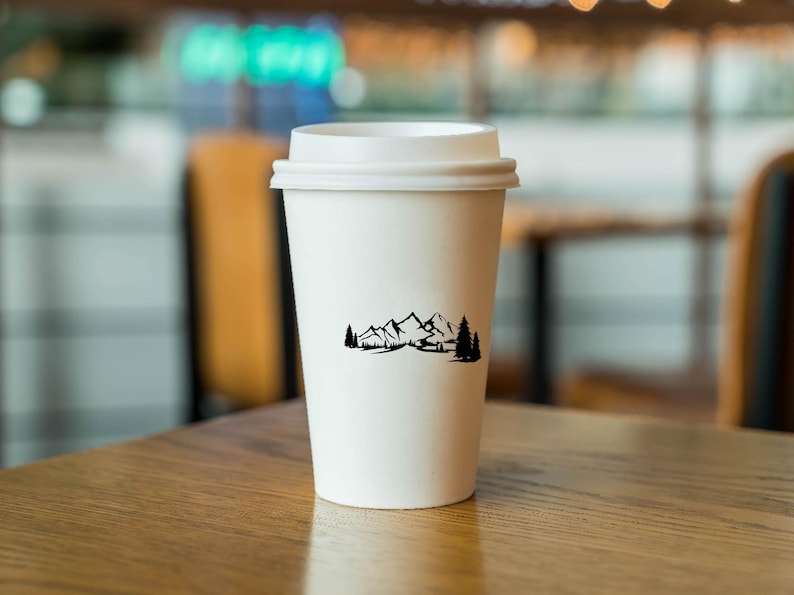 May include: White to-go coffee cup with a black mountain range and trees design. The cup is sitting on a wooden table.