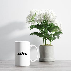 May include: White ceramic mug with a black mountain range design. The mug is sitting on a wooden table next to a potted white flower.